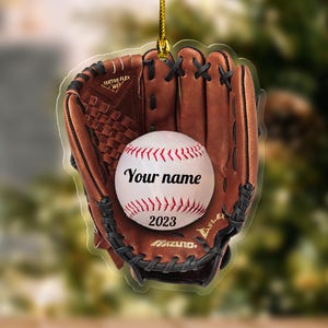 May include: A baseball-themed ornament featuring a brown baseball glove holding a white baseball with red stitching. The baseball has the text "Your name" and "2023". The ornament is suspended by a gold string.