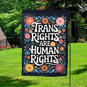 May include: A black garden flag with a floral border and the text "Trans Rights Are Human Rights" in white lettering.