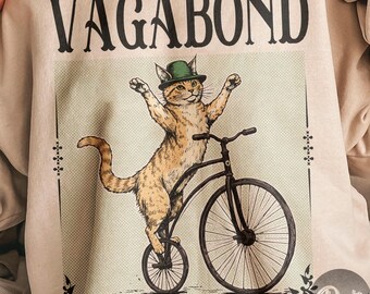 以下が含まれることがあります： ベージュ色のスウェットシャツ。ペニーファージング自転車に乗った猫のグラフィック、緑の帽子をかぶり、上に「VAGABOND」の文字。猫はオレンジと白で、腕を上げています。デザインは装飾的な要素で縁取られています。