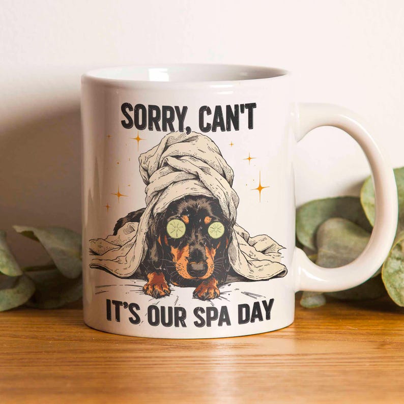 May include: White ceramic mug with a dachshund illustration. The dog is depicted with a towel on its head and cucumber slices on its eyes. The text on the mug reads "SORRY, CAN'T IT'S OUR SPA DAY".