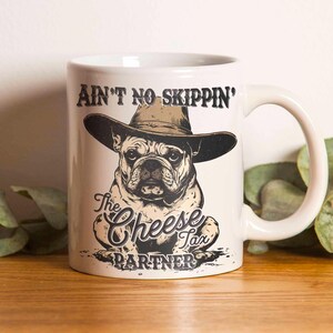 May include: White ceramic mug featuring a bulldog wearing a cowboy hat. The mug has the text "AIN'T NO SKIPPIN'" above the dog and "The Cheese Tax Partner" below. The design is in black and brown tones.