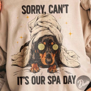 May include: A light beige sweatshirt with the text "SORRY, CAN'T" above an illustration of a dachshund wearing a towel and cucumber slices over its eyes. The text "IT'S OUR SPA DAY" is below the dog.