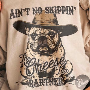 May include: A light beige sweatshirt featuring a bulldog wearing a cowboy hat. The graphic includes the text "AIN'T NO SKIPPIN'" above the dog and "The Cheese Tax Partner" below. The design is in black and brown.