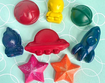 May include: A set of eight space-themed crayons, including a red planet, a green planet, a yellow alien, a red spaceship, a blue spaceship, a blue rocket, a pink star, and an orange star.