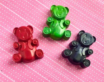 May include: Three small bear-shaped crayons in red, green, and black. The crayons are on a pink background with white polka dots.