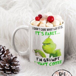 May include: White ceramic mug featuring a green Grinch character graphic. The mug has text that reads "I don't care what day it is. It's early... I'm grumpy I want coffee". The mug is filled with marshmallows and red cranberries.