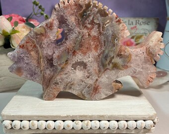 May include: A carved stone fish sculpture in shades of purple, pink, and white, resting on a white wooden stand with decorative beaded trim. The fish has a textured surface and intricate details, showcasing the natural patterns of the stone.