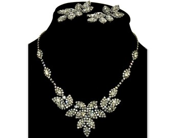 Op de afbeelding: Een sprankelende zilverkleurige ketting en bijpassende oorbellen set. De ketting heeft een bloemmotief met talrijke heldere, ronde en marquise-geslepen strass-steentjes. De oorbellen zijn ook versierd met strass-steentjes in een vergelijkbaar bloemmotief. De sieraden worden gepresenteerd op een zwarte mannequin.