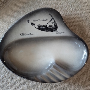 May include: A gray and white ashtray with a map of Nantucket and the words "Atlantic Ocean" and "Nantucket" printed on it. The ashtray has a unique shape and a glossy finish.