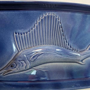 May include: A rectangular ceramic tray in a deep navy blue glaze featuring a raised, embossed relief of a sailfish. The fish is depicted in profile with its prominent dorsal fin extended, swimming across the center of the glossy, smooth surface.