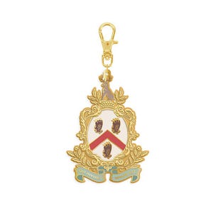 May include: A gold-colored keychain charm with a heraldic crest design. The crest features a white background with a red chevron and three black bear paws. The charm has a gold clasp and is adorned with a small animal figure at the top.