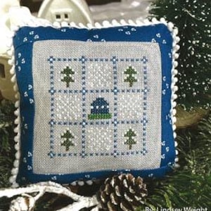 May include: A cross-stitch pattern on a blue pillow with a white background. The pattern is a nine-patch design with a blue and green house in the center and green trees in the corners. The pillow has a white trim with white pom-poms. The text "January Nine Patch" is at the top of the image and "Primrose Cottage Stitches" is at the bottom.