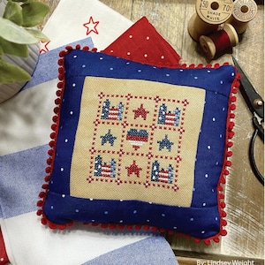May include: A square, patriotic-themed pillow with a blue border and red pom-pom trim. The center features a cross-stitch design with American flags, stars, and a heart. The text "JULY NINE PATCH" and "PRIMROSE COTTAGE STITCHES" are visible.