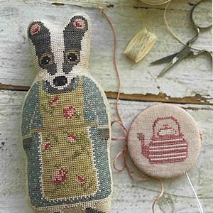 May include: Cross-stitch pattern featuring a badger wearing a green and yellow apron with pink flowers. The badger is holding a teapot in its hands. The pattern is on a white background with the text "Cross-Stitch Pattern" at the bottom.