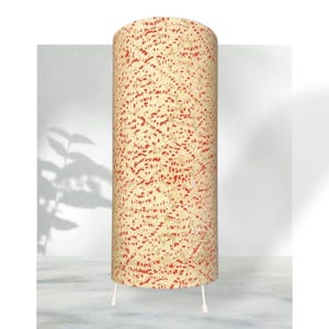 Może przedstawiać: Cylindryczna lampa podłogowa z kremowym kloszem z czerwonym, nakrapianym wzorem. Lampa stoi na trzech białych nogach. Design jest nowoczesny i dekoracyjny, odpowiedni do oświetlenia wnętrz. Wysokość ok. 120 cm.