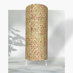 May include: A cylindrical floor lamp with a patterned shade in shades of olive green, beige, and red. The lamp stands on three white legs. The lamp's design features an abstract pattern, adding a decorative element to the room.