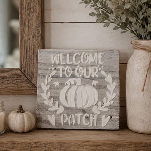 May include: A square wooden sign with a weathered grey finish featuring white painted text that reads WELCOME TO OUR PATCH and a central pumpkin illustration surrounded by leafy vines and small heart shapes, displayed on a wooden surface.