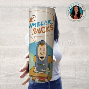 May include: A white stainless steel tumbler with a cartoon character wearing a grey hoodie and an AC/DC t-shirt. The tumbler has a black and white speech bubble that says "Your Tumbler Sucks".