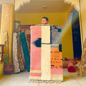 May include: A man holds up a colorful woven rug with geometric patterns in pink, white, black, and orange. The rug is hanging in a doorway with other rugs behind it.