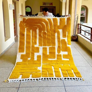 May include: A rectangular, handmade rug with a bold, abstract design in yellow and white. The rug features a geometric pattern with thick, vertical and horizontal lines. The rug has a white fringe along the bottom edge. The rug is being held up by a person.