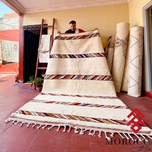 Puede incluir: Un hombre sostiene una gran alfombra blanca tejida a mano con rayas de colores. La alfombra tiene flecos en la parte inferior y est&aacute; extendida sobre un suelo de baldosas. La alfombra est&aacute; rodeada de otras alfombras enrolladas con patrones geom&eacute;tricos.
