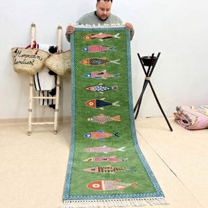 Könnte beinhalten: Ein grüner Läufer mit Fischmuster. Der Teppich hat einen türkisfarbenen Rand und weiße Fransen. Die Fische sind in verschiedenen Farben, darunter Rot, Blau und Orange. Der Teppich wird von einer Person gehalten.