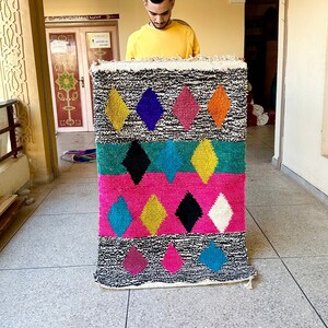 K&ouml;nnte beinhalten: Ein bunter marokkanischer Teppich mit einem geometrischen Diamantenmuster in den Farben Rosa, Blau, Gelb, Gr&uuml;n und Schwarz. Der Teppich wird von einer Person gehalten.