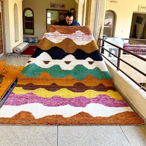 May include: A colorful, hand-woven rug with a geometric pattern in shades of brown, yellow, green, pink, and white. The rug is folded in half and being held by a person.