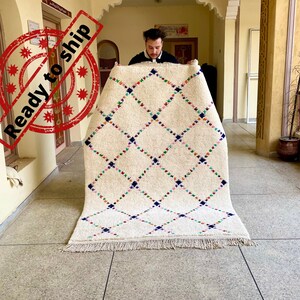 Puede incluir: Una alfombra marroquí blanca con un patrón de diamantes en tonos de azul, rosa, verde y amarillo. La alfombra tiene un borde de flecos y está siendo sostenida por una persona.