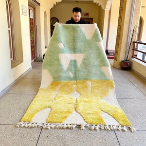 May include: A large, hand-woven rug with a geometric design in shades of green, yellow, and white. The rug has a fringe border and is being held up by a person.
