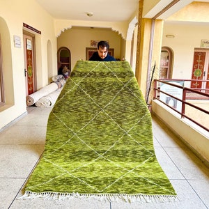 Peut inclure: Un tapis marocain vert et blanc à motifs en losanges est tenu en l'air par une personne dans un couloir. Le tapis a une frange en bas.