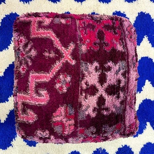 Pu&ograve; includere: Un pouf quadrato e morbido nei toni del magenta scuro, fucsia e rosa chiaro. La superficie strutturata presenta motivi geometrici e floreali. Il pouf &egrave; su un tappeto a zig-zag blu e bianco.