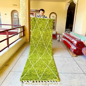 May include: A long, green rug with a diamond pattern and white fringe. The rug is folded in half and is being held by a person.