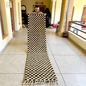 May include: A long, rectangular rug with a checkered pattern in brown and cream colors. The rug has fringe on the ends.