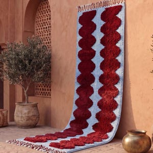 Peut inclure: Un long tapis rectangulaire de type chemin de table, doté d'une base bleu clair avec deux colonnes verticales ondulées en poils longs rouge foncé. Le tapis est terminé par des franges courtes multicolores aux deux extrémités.
