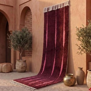 Peut inclure: Un long tapis rectangulaire à la texture hirsute rouge foncé et bordeaux, orné de rayures verticales dans une teinte rose plus claire. Le tapis présente des franges de couleur crème aux extrémités et est suspendu contre un mur en terre cuite.