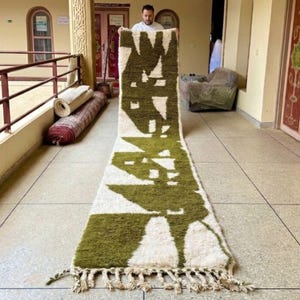 Peut inclure: Un long tapis rectangulaire avec un motif géométrique vert olive et blanc. Le tapis a un bord à franges et est exposé sur un sol carrelé. Le motif du tapis présente des formes et des lignes abstraites, créant une esthétique moderne.
