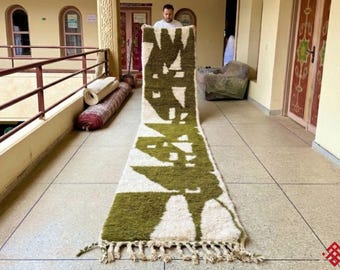 May include: A long, rectangular rug with a geometric design in olive green and white. The rug has a fringed edge and is displayed on a tiled floor. The rug's pattern features abstract shapes and lines, creating a modern aesthetic.
