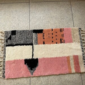 May include: A rectangular rug with a geometric pattern in black, white, pink, and beige. The rug has a fringe border.