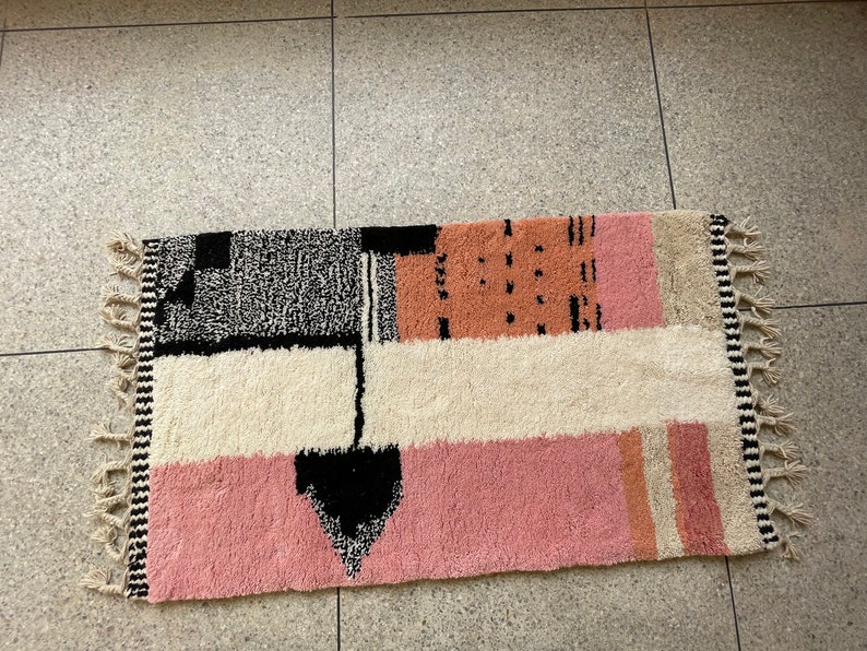 May include: A rectangular rug with a geometric pattern in black, white, pink, and beige. The rug has a fringe border.