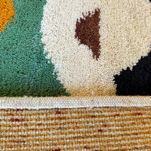 May include: Close-up of a colorful rug with a white, green, brown, black, and yellow geometric pattern. The rug has a white fringe border and a woven backing.