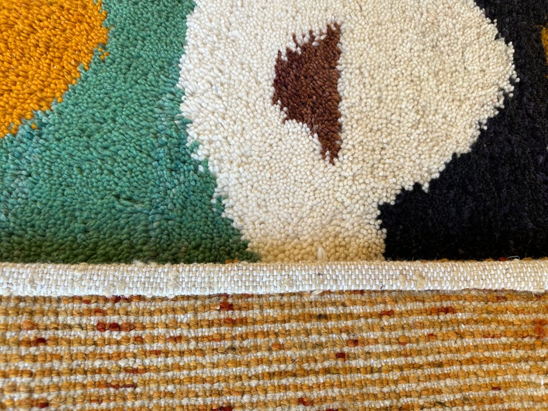 May include: Close-up of a colorful rug with a white, green, brown, black, and yellow geometric pattern. The rug has a white fringe border and a woven backing.