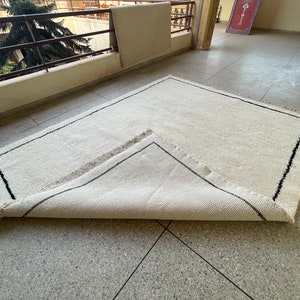 May include: A white rug with a black border and fringe. The rug is folded in half and lying on a tiled floor.