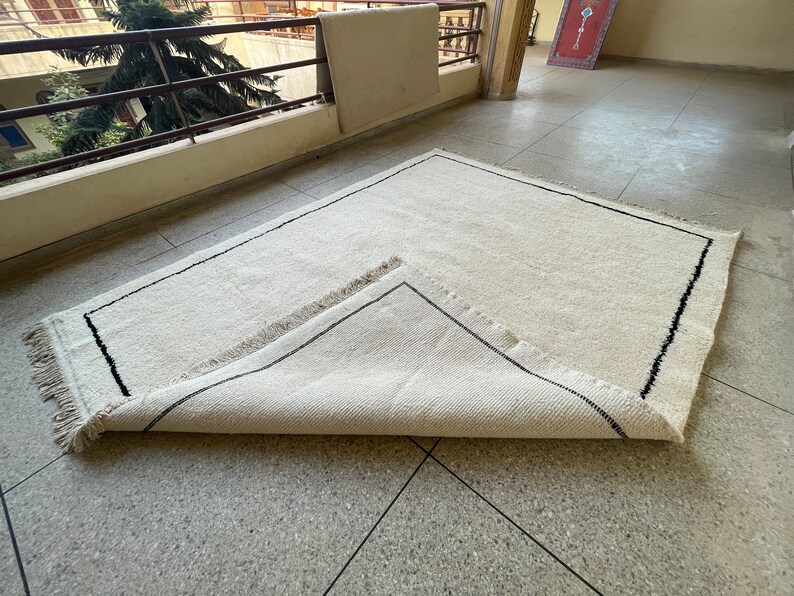 May include: A white rug with a black border and fringe. The rug is folded in half and lying on a tiled floor.