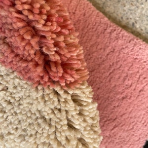 May include: Close-up of a pink, white, and black rug with a fluffy texture. The rug is made of different colored yarns woven together to create a geometric pattern.