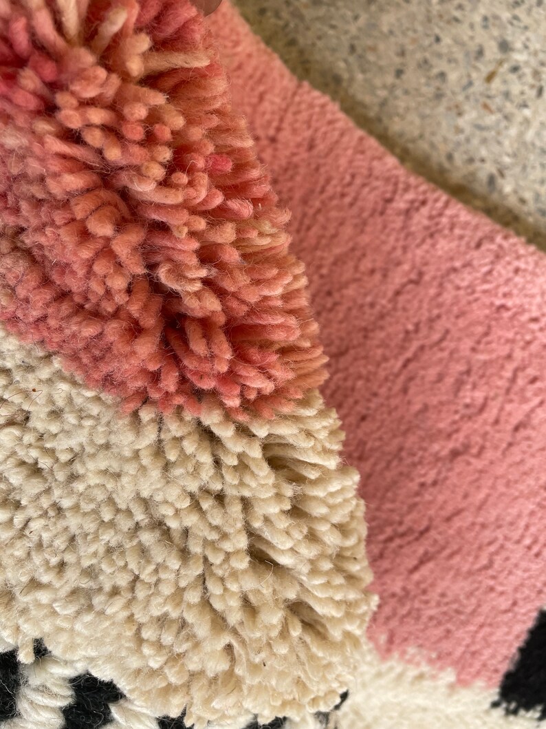 May include: Close-up of a pink, white, and black rug with a fluffy texture. The rug is made of different colored yarns woven together to create a geometric pattern.