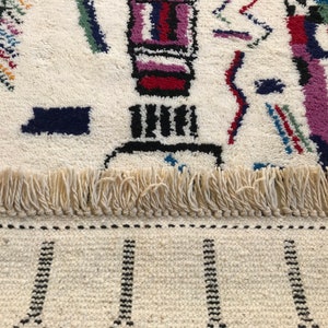 May include: A white rug with a colorful geometric pattern. The rug has a fringe border and a black and white striped pattern at the bottom.