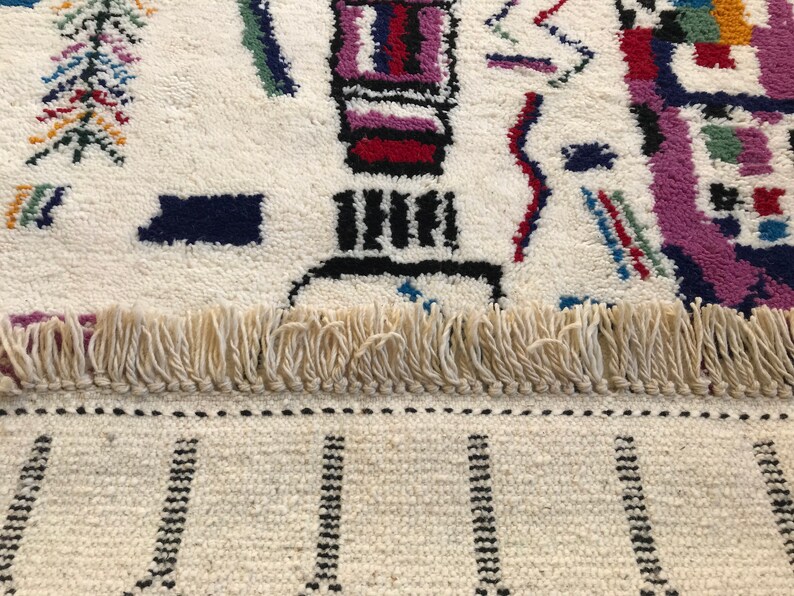 May include: A white rug with a colorful geometric pattern. The rug has a fringe border and a black and white striped pattern at the bottom.