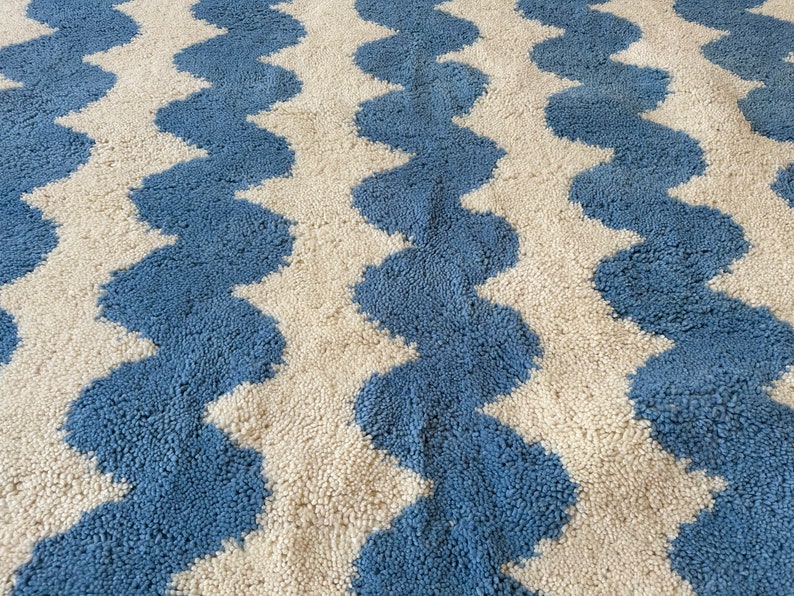 Pu&ograve; includere: Primo piano di un tappeto con un motivo ondulato ripetuto. Il tappeto presenta bande alternate di blu e bianco sporco. La trama appare morbida e soffice, con un pelo alto. Il design &egrave; un motivo geometrico semplice ma audace.