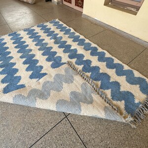 Pu&ograve; includere: Un tappeto rettangolare con sfondo color crema e un motivo ripetuto di zigzag blu. Il tappeto ha un bordo con frange ed &egrave; parzialmente arrotolato, rivelando il retro.